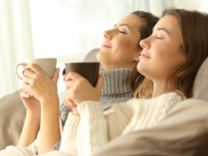 Two women sit comfortably on a couch. The slight smiles and relaxed eyes exude bliss. Each holds a comforting and warm mug filled with something warm and delicious.